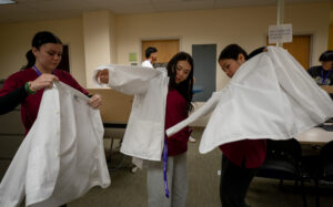 Castle High School students Hailey Faustina, left, Rylie Matsuda, center, and Sophia Morrison don white coats at an educational event and competition at JABSOM April 7, 2026. The competition featured virtual cadaver tables created by Anatomage, a Santa Clara company, who sponsored this program with the Dept of Education and the medical school. . (Craig Fujii/Civil Beat/2026)