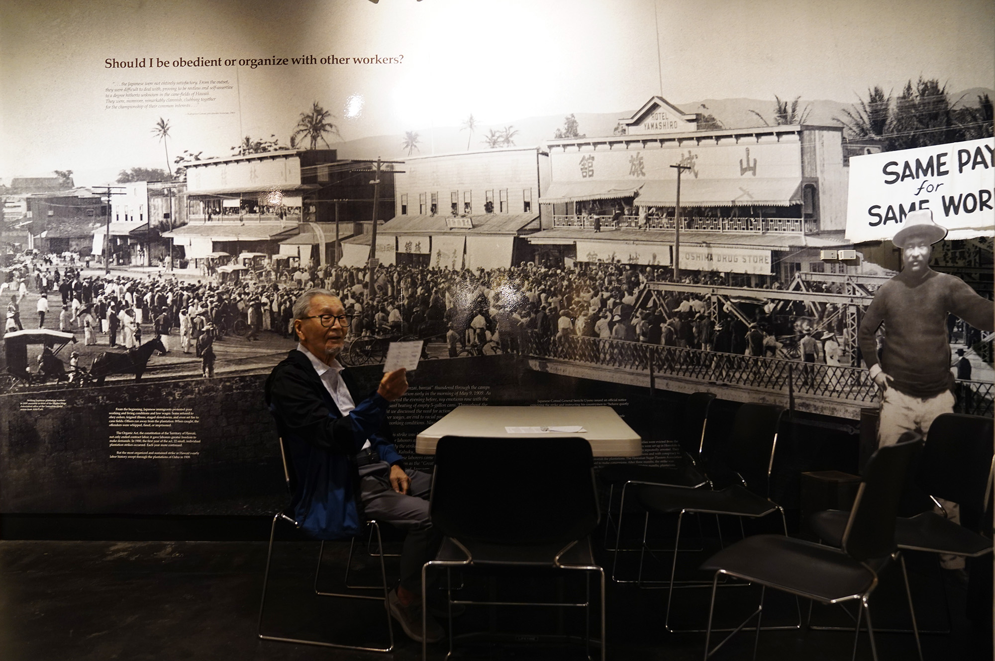 Kazuo Fujii talks story about his relatives unfamiliar to me Thursday, April 9, 2026, at the Japanese Culture Center of Hawaiʻi in Honolulu. Behind him is a large photograph of the Japanese laborer strike of 1909 in which the Hotel Yamashiro served as its headquarters. (Kevin Fujii/Civil Beat/2026)