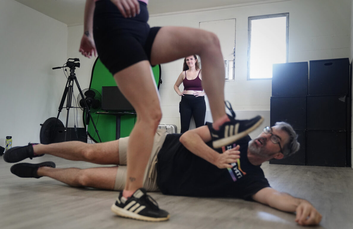 Unarmed stage combat instructor Jasmine Haley Anderson, watches student Miriam Yazdanpanahi kick teaching assistant Andrew Simmons during a choreographed scene Saturday, April 11, 2026, at Manoa Valley Theatre in Honolulu. Anderson stands where the audience would sit. Yazdanpanahi’s foot appears to kick Simmons’ head, but in reality is about two feet away. (Kevin Fujii/Civil Beat/2026)