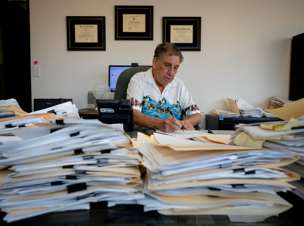A man with graying hair and a white aloha shirt sits at a desk writing with piles of folders in front of him on a desk.