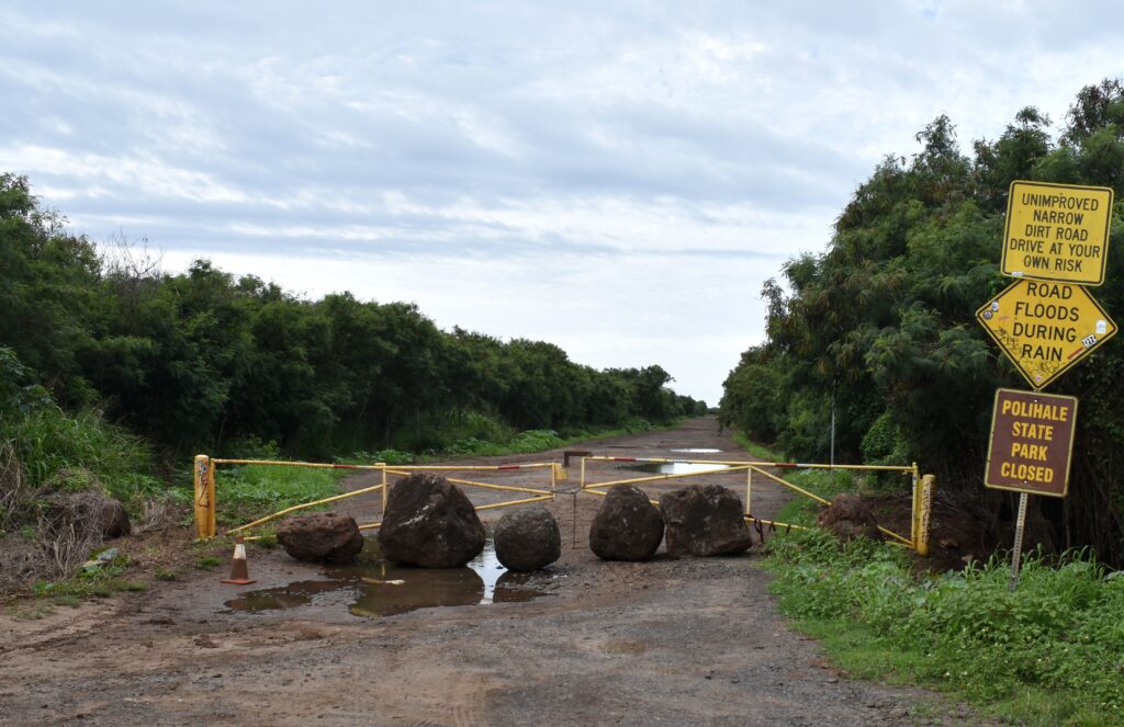 Boulders sit in front of a locked yellow gate, blocking a dirt access road. Yellow and brown signs are to the gate's right, saying "unimproved narrow dirt road drive at your own risk," "road floods during rain," and "Polihale state park closed."