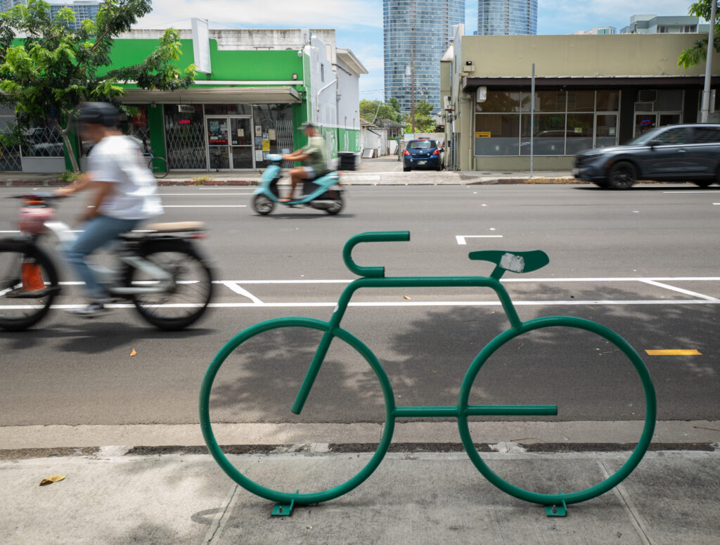 A bicyclist and a moped speed past a bike locking stand on South King Street April 20, 2026. (Craig Fujii/Civil Beat/2026)