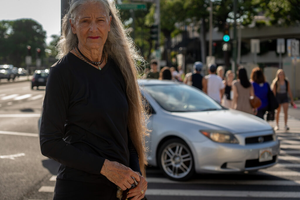 A woman with long graying hair stands next to an intersection where a silver car is in the middle of a crosswalk with several pedestrians out-of-focus in the background.