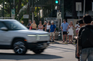 Pedestrians with the right-of-way wait for a turn vehicle to cross over a crosswalk. The light-colored vehicle is blurred by movement.