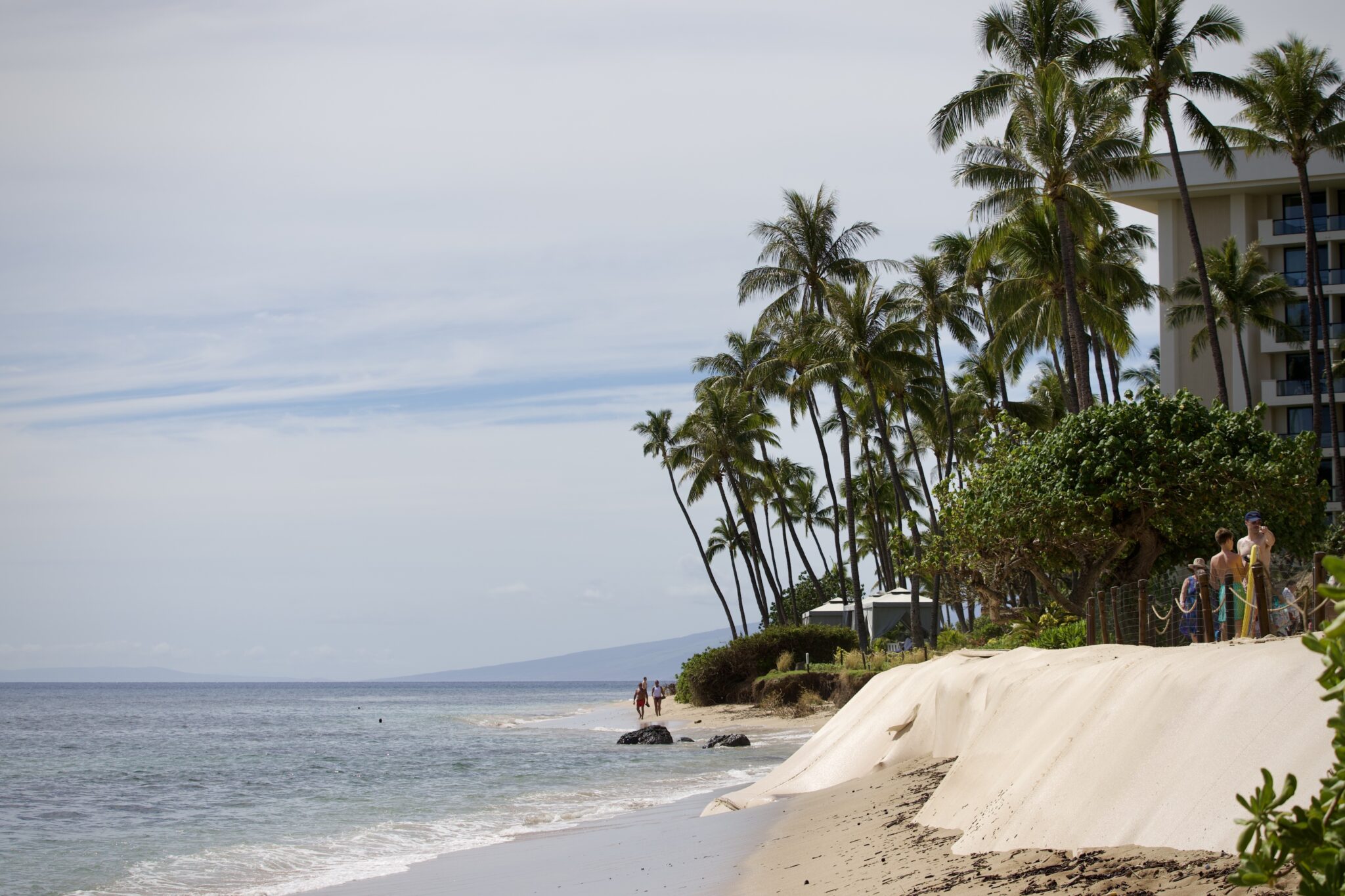 A beige plastic sheet separates a paved pathway and a narrow stretch of Kāʻanapali Beach. Waves lap against the bottom of the barrier.