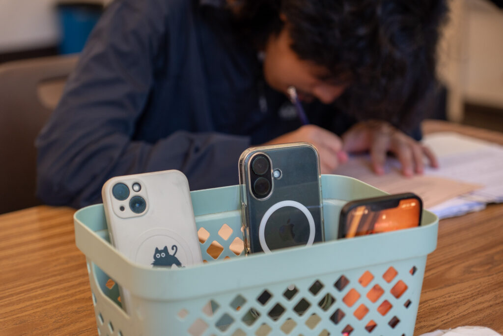 Mobile phones sit in a light blue basket as a student writes with a pencil.