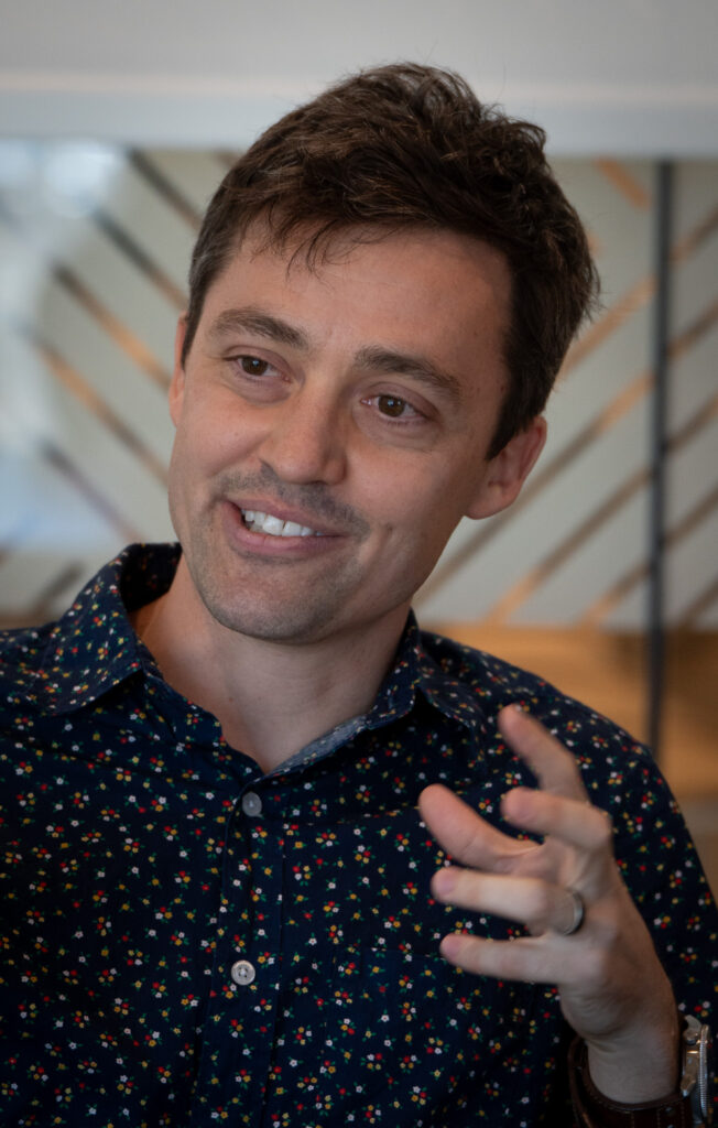 A light-skinned man with dark hair gestures with his left hand. He has a crooked half-smile on his face and wears a dark patterned shirt.
