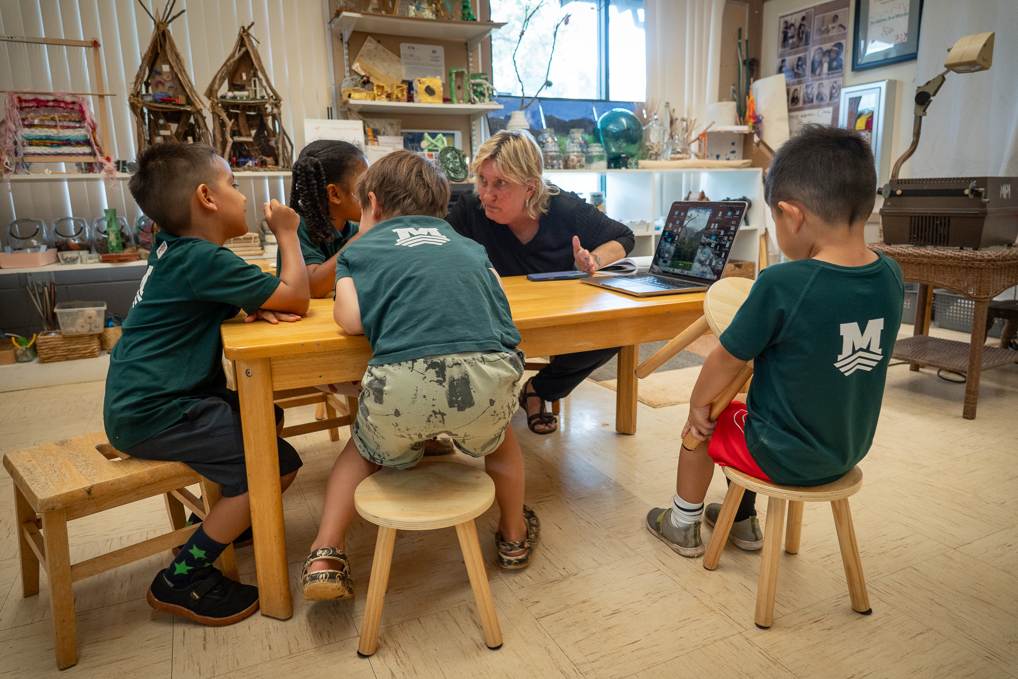 A group of 5-year-old wearing green t-shirts sit at a low wooden table with their teacher and laptop computer.