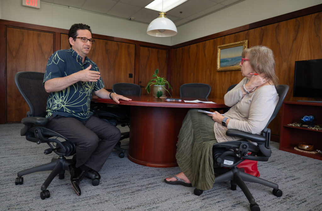 A dark-haired man with glasses and a blue patterned shirt, left, speaks with a woman wearing a beige top and an olive floor-length skirt as they sit at a table in an office.