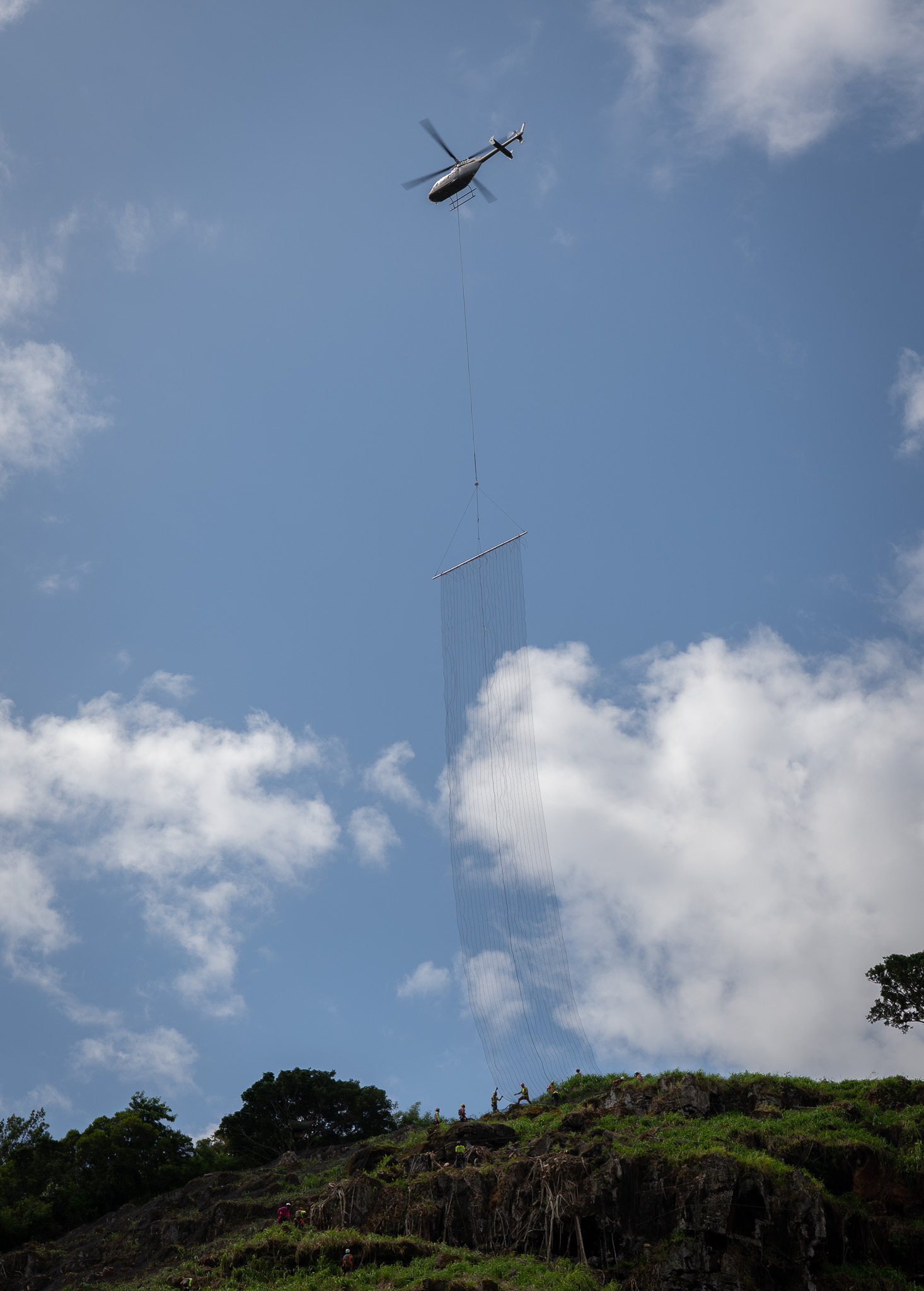 A helicopter lowers a 24 by 150 foot length of rockfall netting next to workers visible on the ridgeline above Waimea Bay May 17, 2026. The 150-foot-long length of netting weighs about 2000 pounds. (Craig Fujii/Civil Beat/2026)