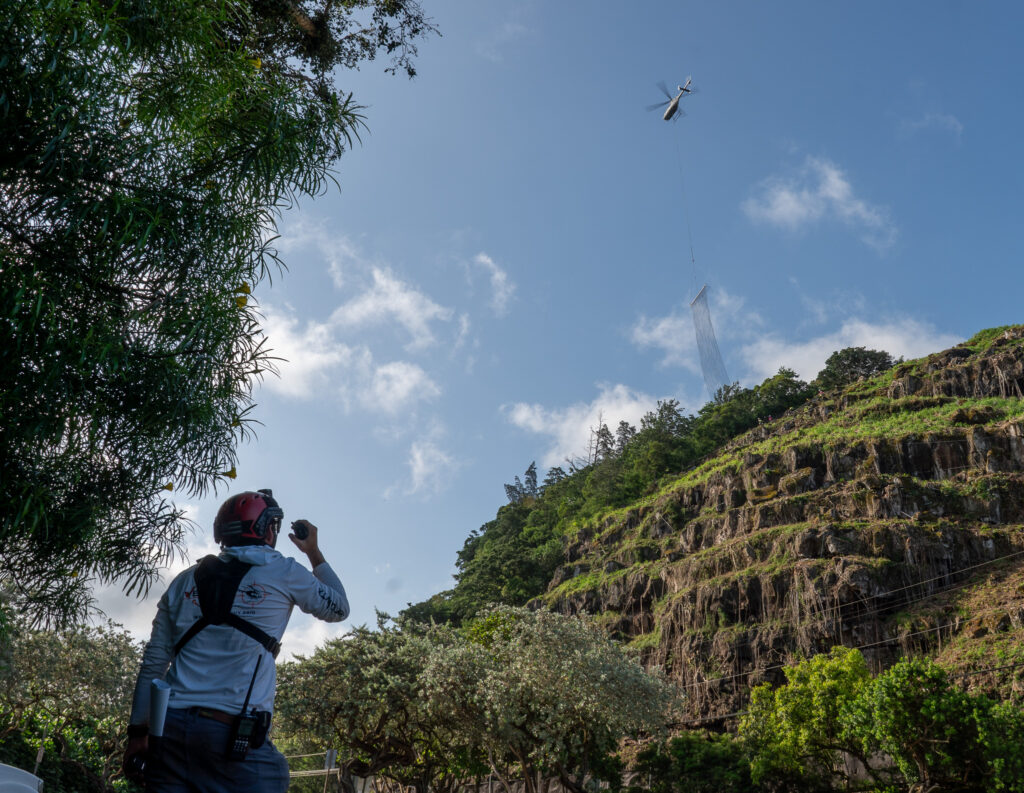 A Vertical Worx (cq) safety worker monitors radio communications as their helicopter lowers a 1000 lb. length of netting onto the unstable hillside above Waimea Bay May 17, 2026. The highway was closed to traffic during operations Sunday. (Craig Fujii/Civil Beat/2026)