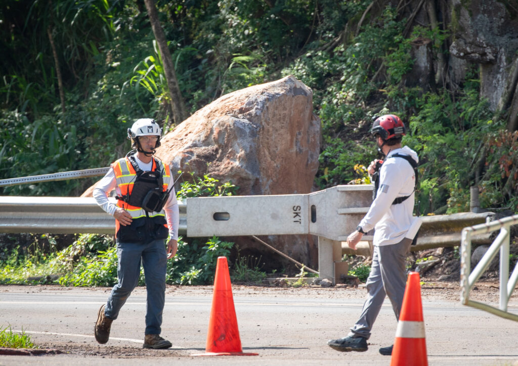 Safety workers with a helicopter crew walk near one of the boulders that fell onto the Kamemehameha Highway from a 230-foot-tall hill above the roadway in early April. The Roadway was closed May 17, 2026 for hillside stabilization. (Craig Fujii/Civil Beat/2026)