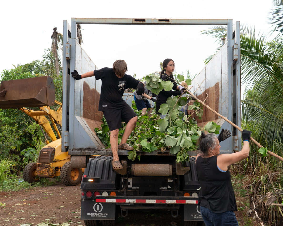 Markus Searls, left, steps down from a roll-off dumpster as he works alongside Lauren Taniyama at Mill 6 Farms in Waialua May 23, 2026. The two worked with volunteers to clear ditches of overgrown trees and plants to improve water flow after the Kona low storms flooded the area. (Craig Fujii/Civil Beat/2026)